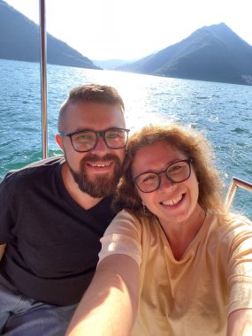 Mutlu çift akıllı telefondan selfie çekiyor ve arka planda çarpıcı dağ manzarası olan manzaralı bir tekne turunun tadını çıkarıyor. Gülümsemeler, suda güzel bir maceranın neşesini ve özgürlüğünü yansıtır.