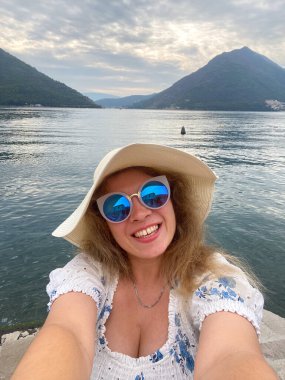 Güneş şapkalı ve güneş gözlüklü komik kadın, arka planda görkemli dağlar olan sakin bir gölün kenarında neşeyle gülümsüyor. Tatili sırasında mutluluk ve doğal güzellikle selfie çekiyordu..