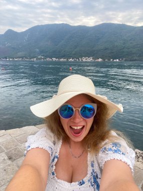 Güneş şapkalı ve güneş gözlüklü komik kadın, arka planda görkemli dağlar olan sakin bir gölün kenarında neşeyle gülümsüyor. Tatili sırasında mutluluk ve doğal güzellikle selfie çekiyordu..