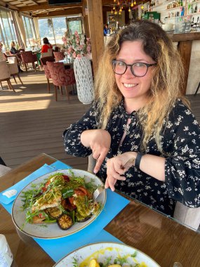 Kadın deniz kenarındaki bir restoranda bir tabak ızgara sebze ve kızarmış tavukla yemek yemekten hoşlanıyor. Parlak ve açık bir atmosfer, palmiye ağaçları ve dışarıdaki sahilin manzarası ile çevrili..