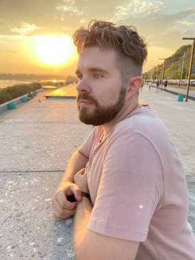 Sakallı adam gün batımına özenle bakar, manzaralı bir korkuluğa yaslanır. Batan güneşin yumuşak parıltısı huzurlu ve yansıtıcı bir atmosfer yaratır..