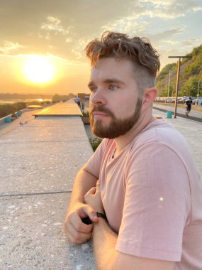 Sakallı adam gün batımına özenle bakar, manzaralı bir korkuluğa yaslanır. Batan güneşin yumuşak parıltısı huzurlu ve yansıtıcı bir atmosfer yaratır..