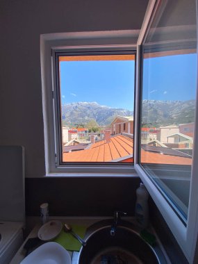 Açık bir pencere ile mükemmel çerçevelenmiş uzak dağların manzarası çatıları ve açık mavi gökyüzünü ortaya çıkarıyor. İçerideki lavabo alanı, dışarıdaki canlı manzarayla çelişiyor.