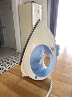 Beyaz ve mavi ütü modern bir mutfak ortamında ahşap bir masanın üzerinde ayakta duruyor ve ev işlerinin özünü yakalıyor. Doğal ışık desenli zemine karşı günlük cihazın parlak tasarımı ve işlevselliğini vurgular.