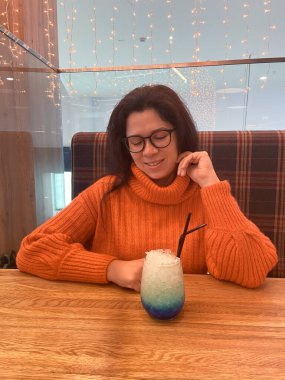 Turuncu kazaklı genç bir kadın bir masada oturuyor, renkli bir içeceğe mutlu bir şekilde bakıyor. Sıcak atmosfer, yumuşak ışıklandırma ve rahat ortam, rahatlama ve keyif hissini arttırır..