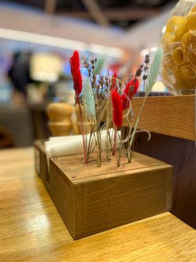 Ahşap bir tutacakta kurumuş çiçeklerin canlı düzenlenmesi, arka planda yanıp sönen ışıklarla samimi bir restoran ortamına renk katar. Kırmızı ve mavinin karışımı sıcak ve yumuşak bir odak noktasında çiçek açar. Şenlikli ve davetkar bir atmosfer yaratır..