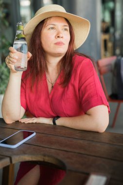 Milenyum kadını hasır şapkalı ve kırmızı elbiseli açık hava kafesinde serinletici bir içki içiyor. Ahşap masa, rahat atmosfer ve rahat poz boş zaman ve yaz keyfi hissi uyandırıyor..