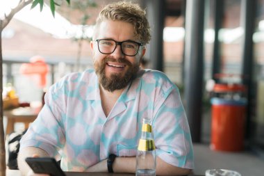 Milenyum neşe dolu gözlüklü ve desenli gömlekli bir adam açık hava kafesinde otururken sıcak bir şekilde gülümsüyor. Gündelik ortam ve parlak ifade pozitifliği ve gevşemiş bir yaz havasını iletir.