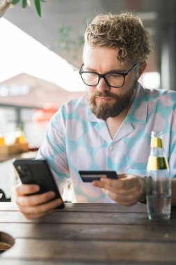 Gözlüklü bir adam açık hava yazlık bir kafede otururken akıllı telefonunu kullanır ve online alışveriş kartı öder. Modern teknoloji, internet alışverişi ve dijital ödemelerin kolaylığı