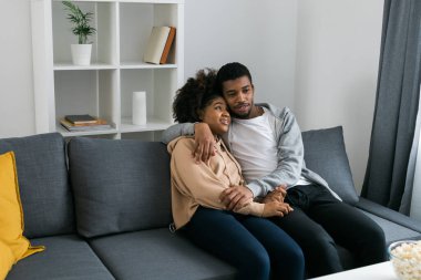 Afro-Amerikan çift rahat gri bir koltukta oturup birbirlerini kucaklıyor ve evlerinde huzurlu bir anın tadını çıkarıyorlar. Aşk ve samimiyet modern ve davetkar bir iç mekanda rahatlık hissi..