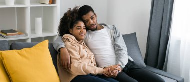 Afro-Amerikan çift rahat gri bir koltukta oturup birbirlerini kucaklıyor ve evlerinde huzurlu bir anın tadını çıkarıyorlar. Aşk ve samimiyet modern ve davetkar bir iç mekanda rahatlık hissi..