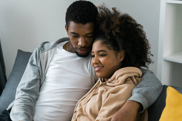 Афроамериканская пара сидит на уютном сером диване, обнимая друг друга и наслаждаясь мирным моментом дома. Любовь и интимность, чувство комфорта в современном и привлекательном интерьере.