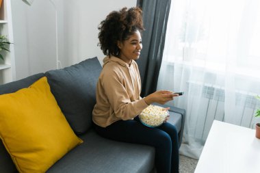 Afrikalı Amerikalı bir kadın gri bir kanepeye oturup film izlerken veya internetten film izlerken bir kase patlamış mısırın tadını çıkarıyor. Modern bir evde rahat ve rahatlatıcı bir eğlence anı. Bağlantı