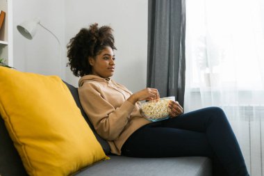 Afrikalı Amerikalı bir kadın gri bir kanepeye oturup film izlerken veya internetten film izlerken bir kase patlamış mısırın tadını çıkarıyor. Modern bir evde rahat ve rahatlatıcı bir eğlence anı. Bağlantı