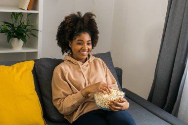 Afrikalı Amerikalı bir kadın gri bir kanepeye oturup film izlerken veya internetten film izlerken bir kase patlamış mısırın tadını çıkarıyor. Modern bir evde rahat ve rahatlatıcı bir eğlence anı. Bağlantı