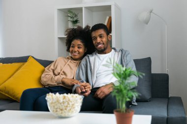 African American couple relaxes on a cozy sofa, sharing a joyful moment while watching a show with a bowl of popcorn. Essence of quality time and comfort in a stylish modern living room. clipart