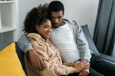 Afro-Amerikan çift rahat gri bir koltukta oturup birbirlerini kucaklıyor ve evlerinde huzurlu bir anın tadını çıkarıyorlar. Aşk ve samimiyet modern ve davetkar bir iç mekanda rahatlık hissi..