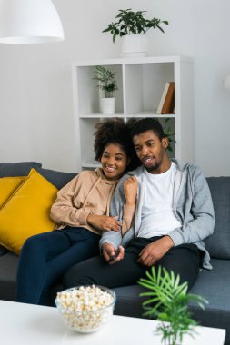 African American couple relaxes on a cozy sofa, sharing a joyful moment while watching a show with a bowl of popcorn. Essence of quality time and comfort in a stylish modern living room. clipart