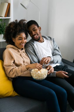 Afro-Amerikalı çift rahat bir kanepede gevşeyip bir kase patlamış mısırla bir programı izlerken keyifli bir anı paylaşıyorlar. Modern bir oturma odasında kaliteli zaman ve konfor esansı..