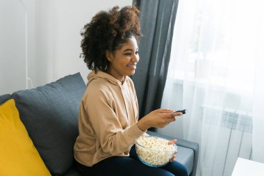 Afrikalı Amerikalı bir kadın gri bir kanepeye oturup film izlerken veya internetten film izlerken bir kase patlamış mısırın tadını çıkarıyor. Modern bir evde rahat ve rahatlatıcı bir eğlence anı. Bağlantı