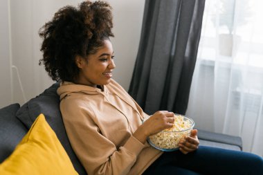 Afrikalı Amerikalı bir kadın gri bir kanepeye oturup film izlerken veya internetten film izlerken bir kase patlamış mısırın tadını çıkarıyor. Modern bir evde rahat ve rahatlatıcı bir eğlence anı. Bağlantı