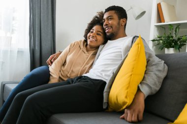 Afro-Amerikalı çift rahat bir kanepede rahatlar, bir dizi izlerken keyifli bir anı paylaşırlar. Modern bir oturma odasında kaliteli zaman ve konfor esansı.