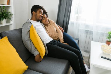 African American couple relaxes on a cozy sofa, sharing a joyful moment while watching a show. Essence of quality time and comfort in a stylish modern living room clipart