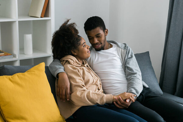 Афроамериканская пара сидит на уютном сером диване, обнимая друг друга и наслаждаясь мирным моментом дома. Любовь и интимность, чувство комфорта в современном и привлекательном интерьере.