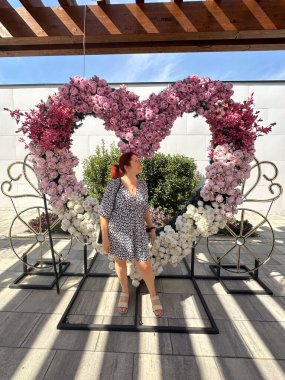 Milenyum kadını, ahşap bir pergola altında pembe ve beyaz güllerle çevrili kalp şeklinde büyük bir çiçek aranjmanının önünde duruyor. Romantizm teması, sevgililer günü ve sanatsal dekorasyon.