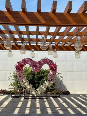 Ampullerle süslenmiş ahşap bir pergola altında kalp şeklinde çiçek aranjmanı romantik ve sanatsal bir açık hava ortamı yaratıyor. Aşk ve Sevgililer Günü konsepti.