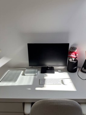 Minimalist beyaz çalışma alanı, bilgisayar monitörü, klavye ve diğer teknolojik aksesuarlar temiz bir masada. Pencereden süzülen parlak güneş ışığı düzenli ve düzenli ayarı vurgular