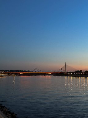 Belgrad 'daki nehrin üzerindeki aydınlık bir köprünün alacakaranlık manzarası sakin sularda huzur yansımaları sergiliyor. Sırbistan 'da kentsel mimarinin ahengi ve akşam karanlığında doğanın solgunluğu.