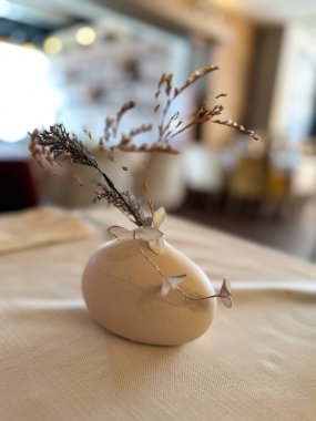 Küçük bir seramik vazo içinde kuru dallar bulunan zarif bir restoran masası. Bulanık arkaplan derinlik ekler ve masa üstü dekorunun basitliğini vurgular.