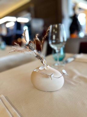 Elegant restaurant table with a small ceramic vase holding dry branches. The blurred background adds depth and highlights the simplicity of the tabletop decor. clipart