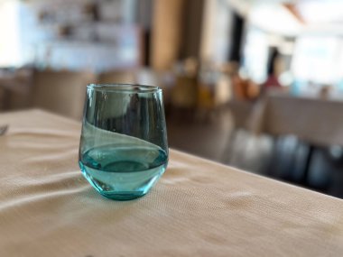 Lokanta masasındaki turkuaz bir cam bardağın yumuşak odak noktası. İnce ışıklandırma ve bulanık arkaplan zarif ve minimalist bir kompozisyon oluşturur.