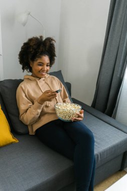 Genç Afrikalı Amerikalı kadın bir televizyon şovunun tadını çıkarırken elinde bir kase patlamış mısır tutuyor. Parlak gülümseme, rahat bir evde mutluluk ve rahatlık yayar..