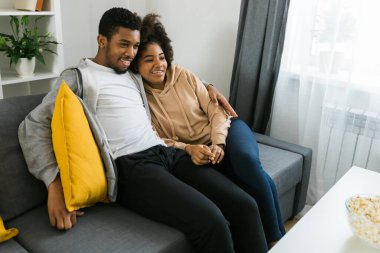 Mutlu Afrikalı Amerikalı çift televizyon izlerken birlikte kaliteli zaman geçirmekten hoşlanıyor. Kanepeye yakın oturan erkek ve kadın rahat bir yuva ortamında rahatlık ve iletişim gösteriyorlar. TV, dizi