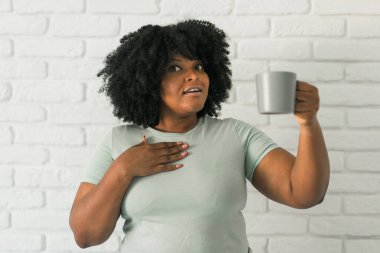 African American surprised woman holding a gray mug while pointing at herself, conveying emotions of shock and curiosity. Moments of self-realization, humor, and unexpected situations in daily life. clipart