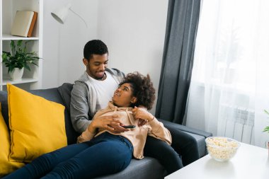 Afro-Amerikalı çift rahat bir kanepede gevşeyip bir kase patlamış mısırla bir programı izlerken keyifli bir anı paylaşıyorlar. Modern bir oturma odasında kaliteli zaman ve konfor esansı..