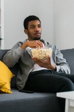 Afro-Amerikalı adam televizyon izlerken ve televizyon izlerken tek başına kanepede oturup patlamış mısırın tadını çıkarıyor..