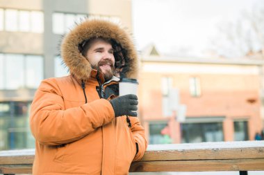 Turuncu parka giymiş gülen adam dışarıda sıcak kahve içmekten hoşlanıyor. Arka planda kış manzarası var. Boşluğu kopyala.