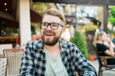 Gözlüklü sakallı adam dışarıda bir kafede otururken neşeyle gülüyor.