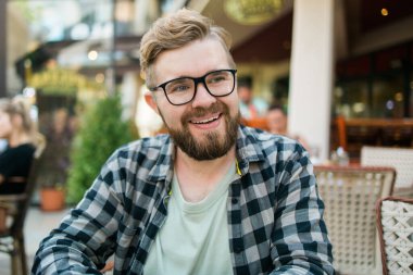 Gözlüklü sakallı adam dışarıda bir kafede otururken neşeyle gülüyor.