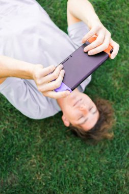Neşeli adam gülümseyip eğlenirken portatif konsolla oyun oynayıp çimlerin üzerinde yatıyordu. Teknoloji ve oyun bağımlılığı kavramı.