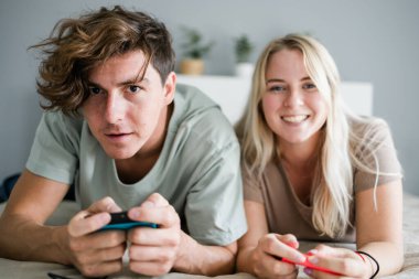 Genç çift yatak odasındaki yatakta yatarken video oyunu oynuyorlar, hafta sonları eğleniyorlar ve eğleniyorlar. Teknoloji ve oyun konsepti.