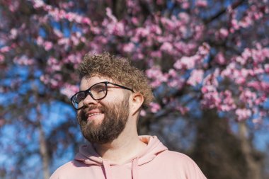 Alerjisi olan bir adam bahar mevsimsel alerjisinden zevk alıyor. Baharda çiçek açan ağacın önünde gülümseyen mutlu sakallı adamın portresi. Bahar çiçekleri ve alerji konsepti. Boşluğu kopyala.