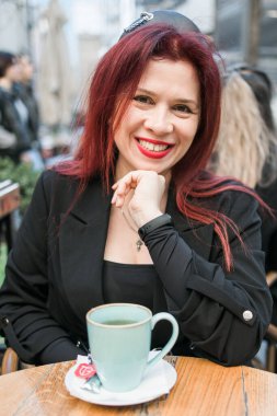 Restoranın terasında parlak kırmızı rujlu güzel bir kadın oturuyor. Kahvede poz veren siyah elbiseli kızıl saçlı harika bir kadın.
