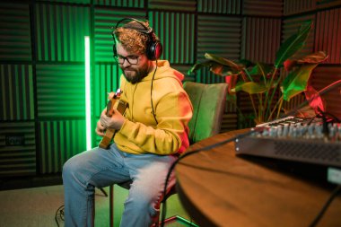 Erkek şarkıcı bir stüdyo mikrofonuyla şarkı kaydediyor ve bir müzik stüdyosunda gitar çalıyor. Müzik sektöründe performans ve gösteri, müzik yayını ve kaydı