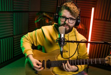 Erkek şarkıcı bir stüdyo mikrofonuyla şarkı kaydediyor ve bir müzik stüdyosunda gitar çalıyor. Müzik sektöründe performans ve gösteri, müzik yayını ve kaydı