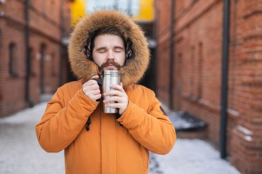 Kışın parka giyen adam termosu kapalı bir şekilde karlı şehir sokağında tutuyor. Soğuk havada sıcaklık, yalnızlık ve konfor.
